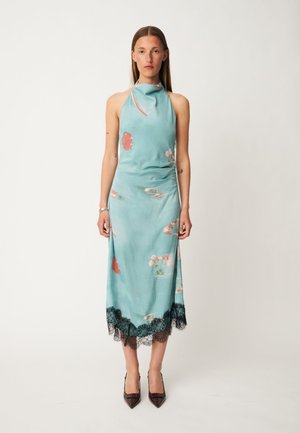Femme portant une robe sans manches bleu clair à fleurs avec un bord en dentelle noire à l'ourlet, debout, les bras détendus le long du corps.