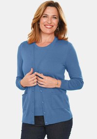 Cardigan bleu en tissu tricot doux avec un col en V, fermeture à boutons, deux poches avant et poignets côtelés. Porté par-dessus un haut intérieur assorti.