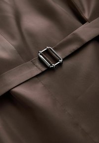 Boucle métallique fixant une ceinture en tissu marron sur un tissu marron lisse, montrant la texture détaillée et les coutures.