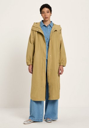 Frau steht und trägt einen langen, hellbraunen Kapuzenmantel über einem blauen Jeanshemd und weiten Jeanshosen mit silbernen flachen Schuhen.