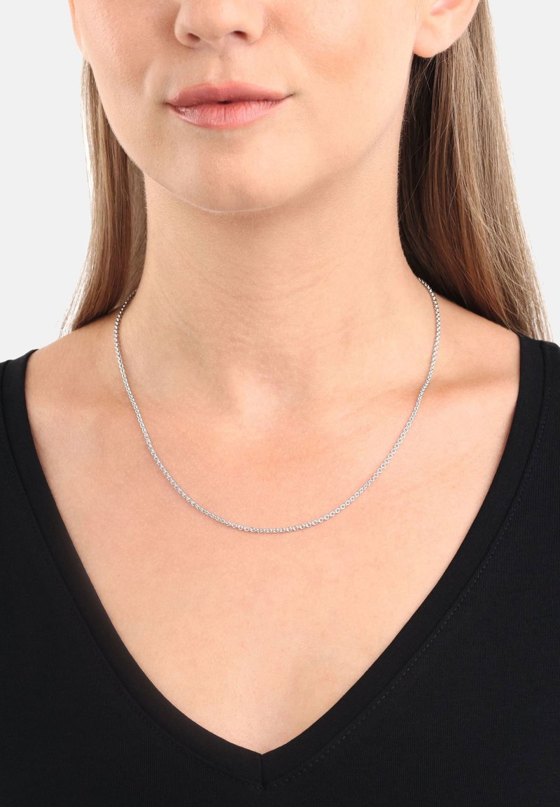 Collar de cadena de plata con un diseño delicado y sutil. Presenta pequeños eslabones entrelazados, llevado por una persona con una blusa negra de escote en V.