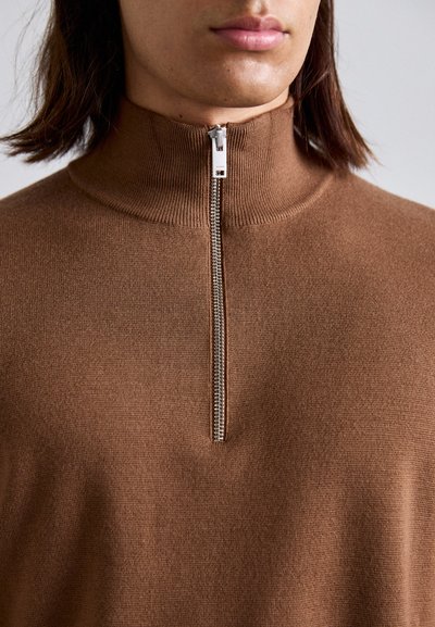 Nahaufnahme einer Person, die einen braunen Pullover mit hohem Kragen trägt, dessen silberner Reißverschluss teilweise geschlossen ist.