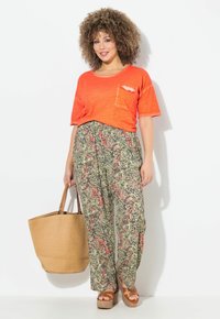 Maglietta cropped arancione con taschino sul petto, pantaloni larghi a fantasia verde e rosso, borsa a tracolla intrecciata color sabbia e sandali in platform marroni.