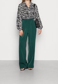 Blouse à fleurs en noir et blanc à manches longues, associée à un pantalon vert à jambes larges. Accessoires : un petit sac à main noir.