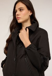 Tânără cu păr lung și brunet, purtând o haină neagră cu nasturi, ținând gulerul cu o mână, privind în lateral.