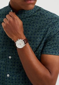 Brun läderremklocka med silverfärgad boett och vit urtavla som visar arabiska siffror. Bäraren har på sig en mönstrad mörkgrön skjorta med geometrisk design.