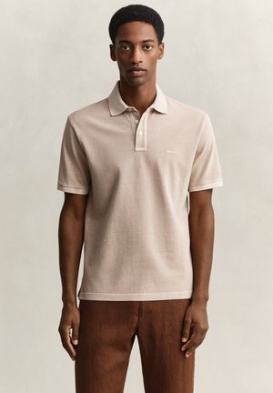 Mann, der nach vorne blickt, trägt ein beiges Polohemd mit kurzen Ärmeln und braune Hose vor einem einfarbigen hellen Hintergrund.