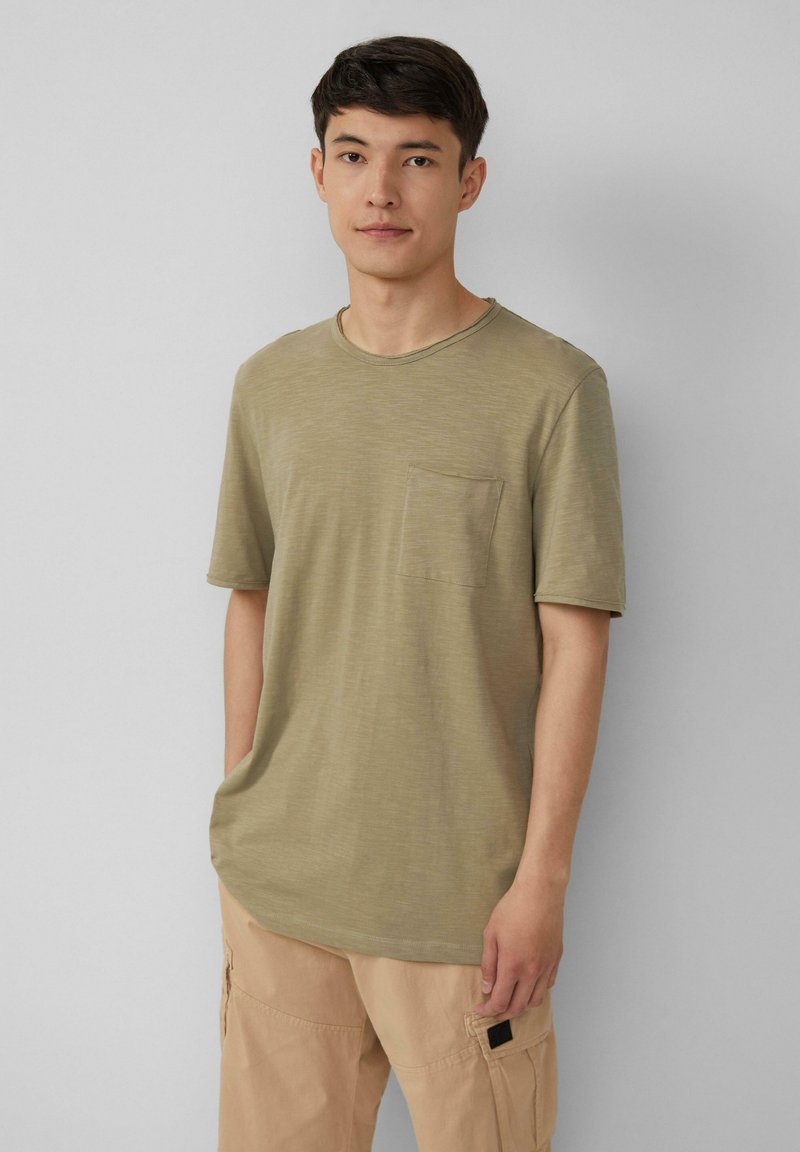 Olijfgroen t-shirt met korte mouwen van zachte, lichte stof, met een ronde hals en een borstzak. Draagt bij beige cargo-broek.