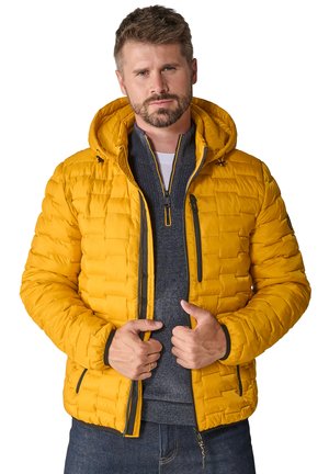 Gelbe Pufferjacke mit Kapuze, gestepptem Design und schwarzen Reißverschlussakzenten. Über einem grauen Pullover getragen, der einen engen Schnitt und gerippte Ärmelbündchen aufweist.