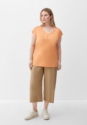 Orange short-sleeve top with a round neck and button detail, paired with tan wide-leg pants and white sneakers. Smooth fabric texture.