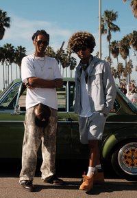 Twee personen staan naast een vintage groene auto. De één draagt een wit T-shirt, grijze broek en een zonnebril; de ander heeft een grijze jas en korte broek aan.