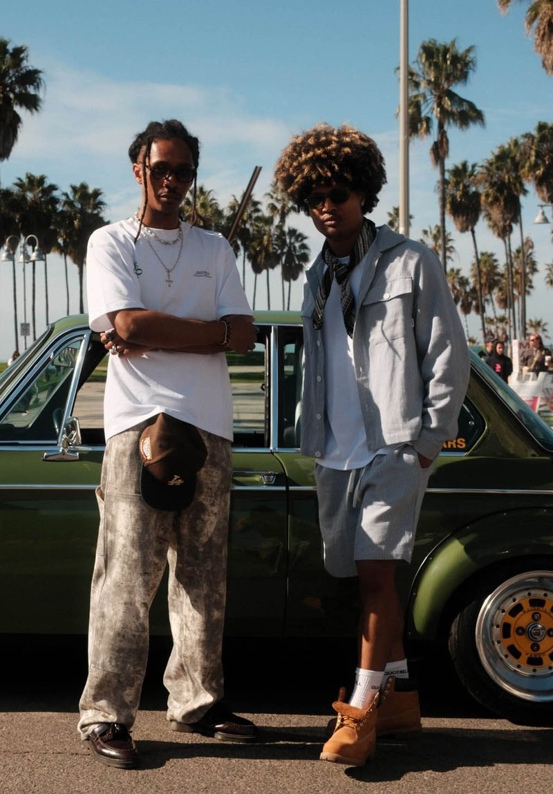 Twee personen staan naast een vintage groene auto. De één draagt een wit T-shirt, grijze broek en een zonnebril; de ander heeft een grijze jas en korte broek aan.