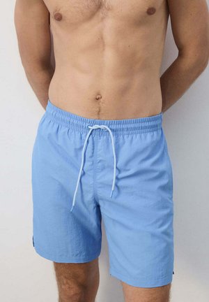 Man wearing light blue swim shorts with white drawstring, standing against plain light wall, hands behind back, torso visible.
