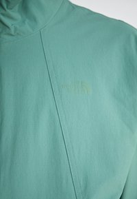 Light teal jacket with a high collar, textured fabric, and a subtle logo. Features diagonal stitching across the front.