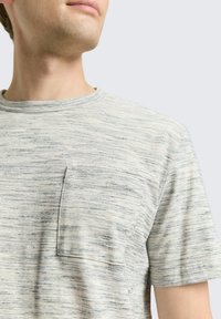 Lichtgrijze t-shirt met een gestructureerd, gestreept patroon, ronde halslijn en een kleine borstzak aan de linkerkant. Korte mouwen.