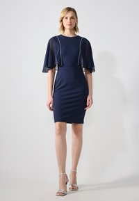 Robe marine avec des manches transparentes et fluides et une silhouette ajustée. Présente un col rond et de subtils embellissements le long du bustier. Talons argentés.