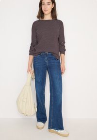 Raidallinen pitkähihaiset paita tumman burgundin ja kermanvärisen yhdistelmänä, yhdistettynä sinisiin leveisiin farkkuihin. Malli pitää kädessään kermanväristä laukkaa ja seisoo valkoista taustaa vasten.