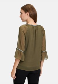 Blusa verde oliva con tessuto trasparente, maniche a campana e rifinitura argento ai bordi delle maniche e al collo. Texture liscia, vestibilità ampia.