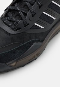 Zapatilla atlética negra con una mezcla de materiales texturizados, cordones redondos y tres franjas blancas de acento. Incluye un logo de adiPrene en la suela.