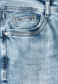 Tissu en denim bleu clair avec un délavage, comprenant une poche avant ornée de clous décoratifs et des détails de couture visibles le long des bords.