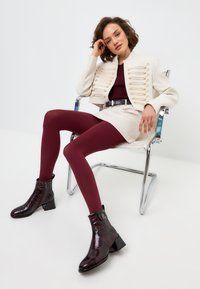 Veste militaire crème avec des boutons décoratifs, leggings bordeaux et bottines noires en cuir verni avec un talon épais. Assise sur une chaise en chrome.