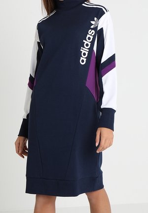 Kobieta nosząca sukienkę dresową Adidas w kolorze granatowym, białym i fioletowym z wysokim kołnierzem i logo na piersi oraz paskami na rękawach.