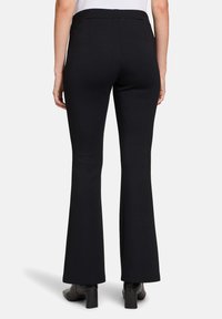 Pantalons évasés noirs au toucher lisse, avec une taille haute, des poches arrière et une coupe ajustée. Portés avec des bottes à talons noires.