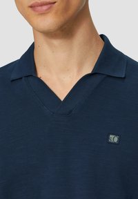 Tumed sinine polo särk V-kaelusega, tekstuurse kangaga ja väike logoga paisutus vasakul rinnal.