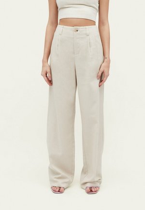 Kvinde iført beige bukser med høj talje og vide ben med knap- og lynlås lukning, kombineret med en hvid cropped top og åbne sandaler.