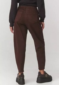 Brązowe spodnie z wysokim stanem i zwężającymi się nogawkami, z dwiema tylnymi kieszeniami i gładką teksturą, połączone z masywnymi czarnymi butami.