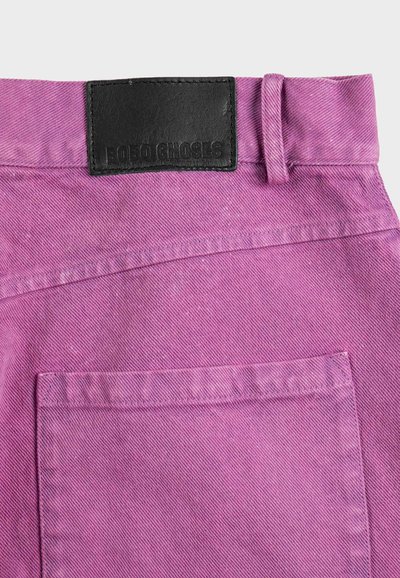 Gros plan sur la poche arrière et la ceinture d'un pantalon en denim violet avec une étiquette en cuir noir cousue, présentant le nom de la marque embossé.