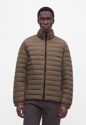 Junger Mann trägt eine braune gesteppte Jacke mit "AX" Logo-Muster und schwarzem Reißverschluss, steht vor einem weißen Hintergrund.