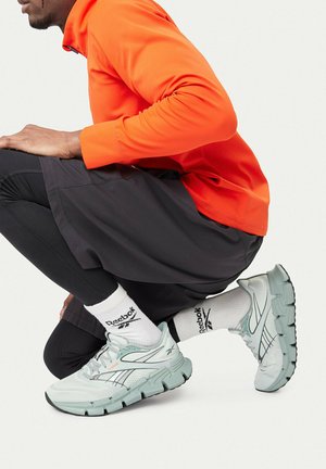 Man wearing white Reebok sneakers, white Reebok socks, black shorts and leggings, and an orange long-sleeve top squatting on one knee.