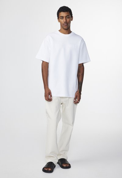 Weißes, kurzärmeliges T-Shirt mit lockerer Passform, kombiniert mit cremefarbenen, lässigen Hosen und schwarzen Slippers. Minimalistisches Design.