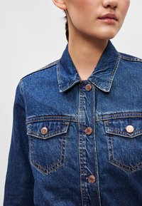 Jeansjakke i mørkeblå, med klassisk krage, kobberfargede knapper og to brystlommer med sømdetaljer. Teksturert stoff.