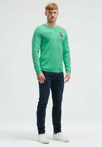 Grünes langärmliges Hemd mit rundem Halsausschnitt, das bedruckte Grafiken und ein Patch aufweist. Kombiniert mit dunklen Jeans und weißen Sneakers.