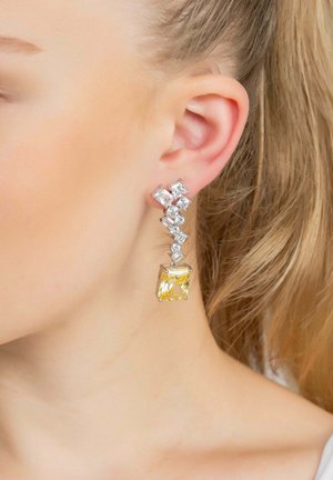Dramatic drop earrings feature a yellow rectangular stone surrounded by clear gemstones in a silver setting, with a zigzag design.