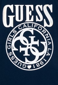 Tume sinine T-särk, millel on valget värvi trükitud tekst "GUESS" ja ümar logo, kus on omavahel põimunud tähed, asutatud 1981, ning väike südame aktsent.