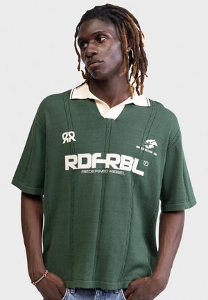 Fiatal férfi dreadlock hajjal, zöld pólót visel, amelyen fehér felirat olvasható: "RDFRBL Redefined Rebel", sima szürke háttér előtt áll.