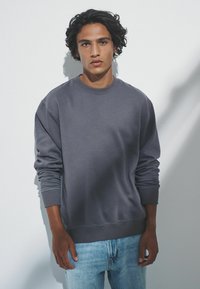 Pelēks kreklu džemperis ar ribotām piedurknēm un apakšmalu. Uzvilkts virs gaiši zilajiem nodiluma džinsiem, kas izceļas ar gludu tekstūru un brīvu piegriezumu.