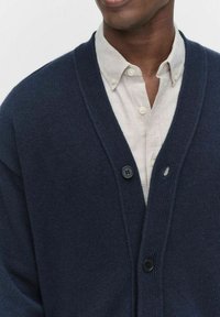 Marinblå cardigan i strukturerad stickad kvalitet, med V-ringning, två svarta knappar och bärs över en ljusfärgad knäppt skjorta.
