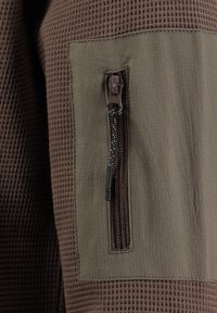 Brązowy materiał o teksturowanym wzorze; zawiera kieszeń na zamek błyskawiczny w kontrastującym zielonym odcieniu. Zamek ma uchwyt z nakrapianym wzorem.