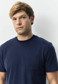 Junger Mann mit hellbraunen Haaren und blauen Augen, der ein marineblaues T-Shirt mit einer Brusttasche trägt, steht vor einem weißen Hintergrund.