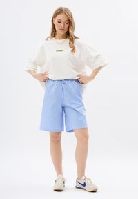 Witte oversized t-shirt met groene tekst, gecombineerd met blauw-wit gestreepte shorts. Model draagt beige sneakers met blauwe accenten.
