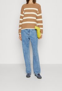 Mujer con suéter de rayas beige y blanco, jeans azules holgados, zapatillas deportivas azules, sosteniendo un bolso verde lima a su lado.