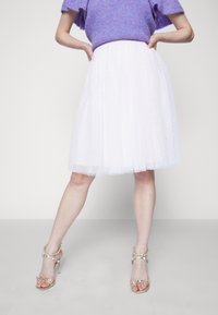 Witte tule midi-jurk met stippenaccenten, gecombineerd met gouden hoge hakken met decoratieve banden. Trui in paarse gebreide stof.