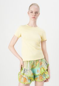 Keltainen lyhythihainen t-paita, istuva malli. Yhdistetty värikkäisiin röyhelöhihaisiin shortseihin, joissa on vihreitä, sinisiä ja oransseja kuvioita sekä lovireunat.