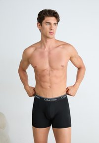 Zwarte katoenen boxershorts met een gladde textuur en een gemerkte tailleband, tentoongesteld op een model zonder shirt, dat een strakke pasvorm benadrukt.