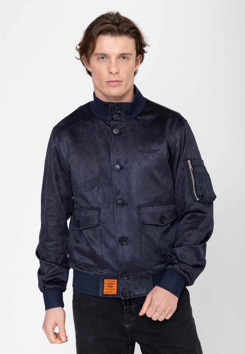 Mann trägt eine navyblaue Bomberjacke mit Knöpfen, zwei Fronttaschen und einer Reißverschlusstasche am linken Ärmel, steht vor einem schlichten Hintergrund.