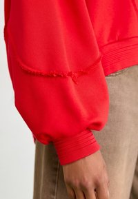 Red sweatshirt featuring puffed sleeves, ribbed cuffs, and a raw hem detail. Soft fabric with visible stitching accents.